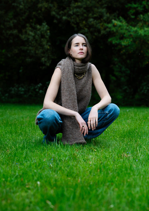 Woman sitting on grass wearing a knitted brown scarf, knitted brown sleeveless top and blue jeans.