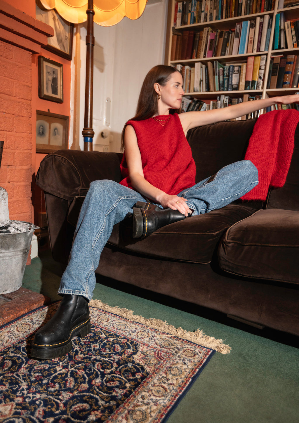 Woman in red knit sleeveless top, blue jeans and black boots sitting on a brown sofa in a cozy room with bookshelves and a fireplace.