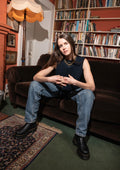 Woman in dark sleeveless knitted top sitting on a brown couch in a room with bookshelves and a lamp.