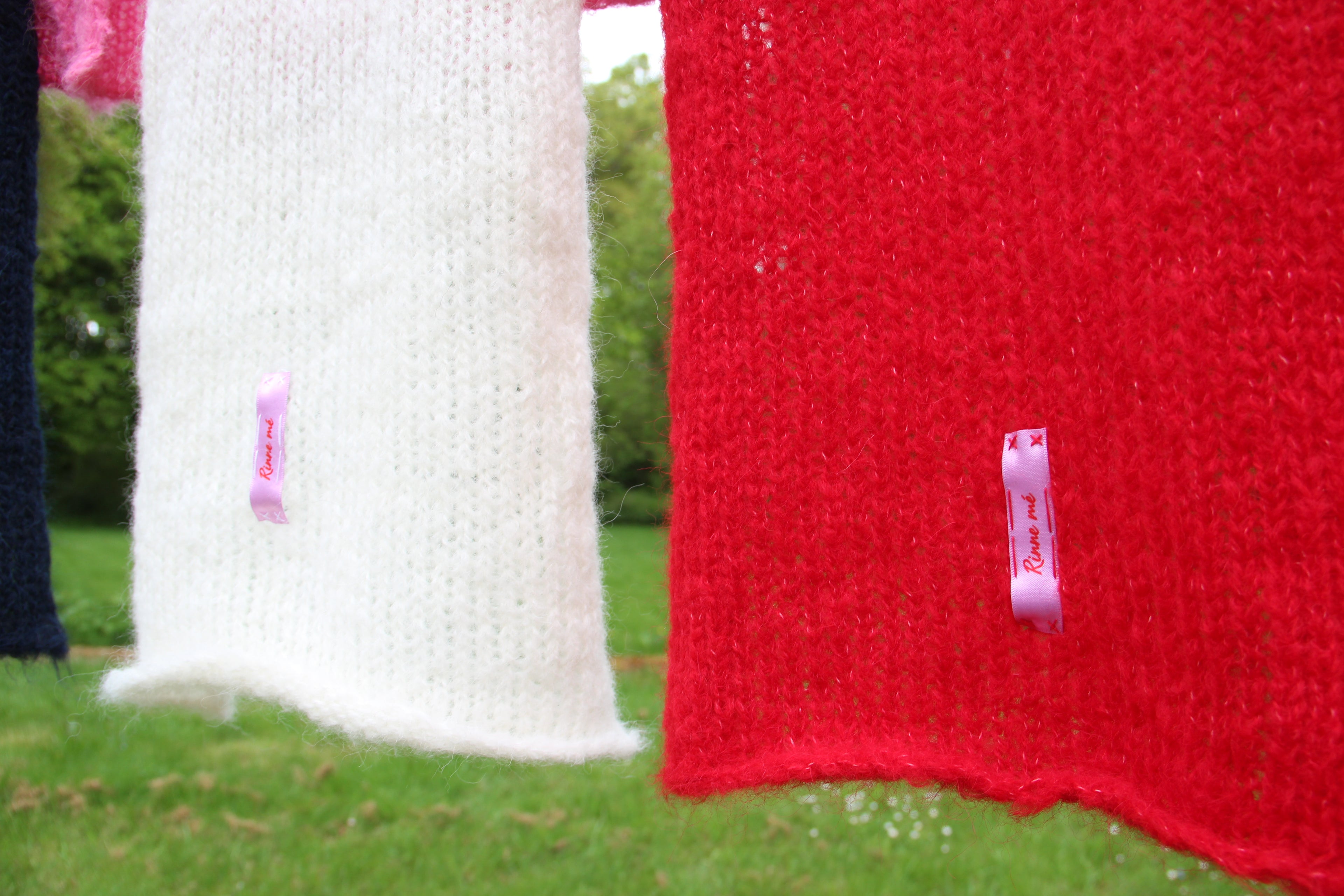 Two knitted scarves hanging outdoors on a line, one white and one bright red, each with a small pink ribbon tag. Grass and greenery are visible in the background.