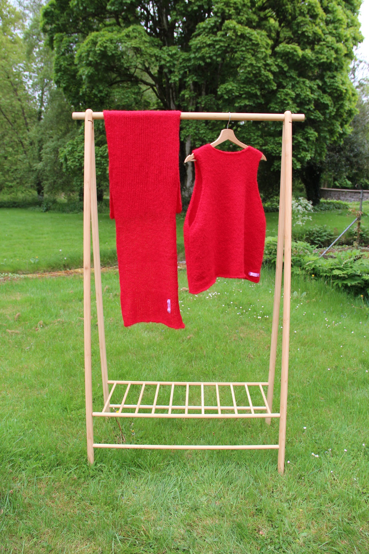 Clothes stand in garden with bright red scarf and red sleeveless sweater hanging on it, trees in background