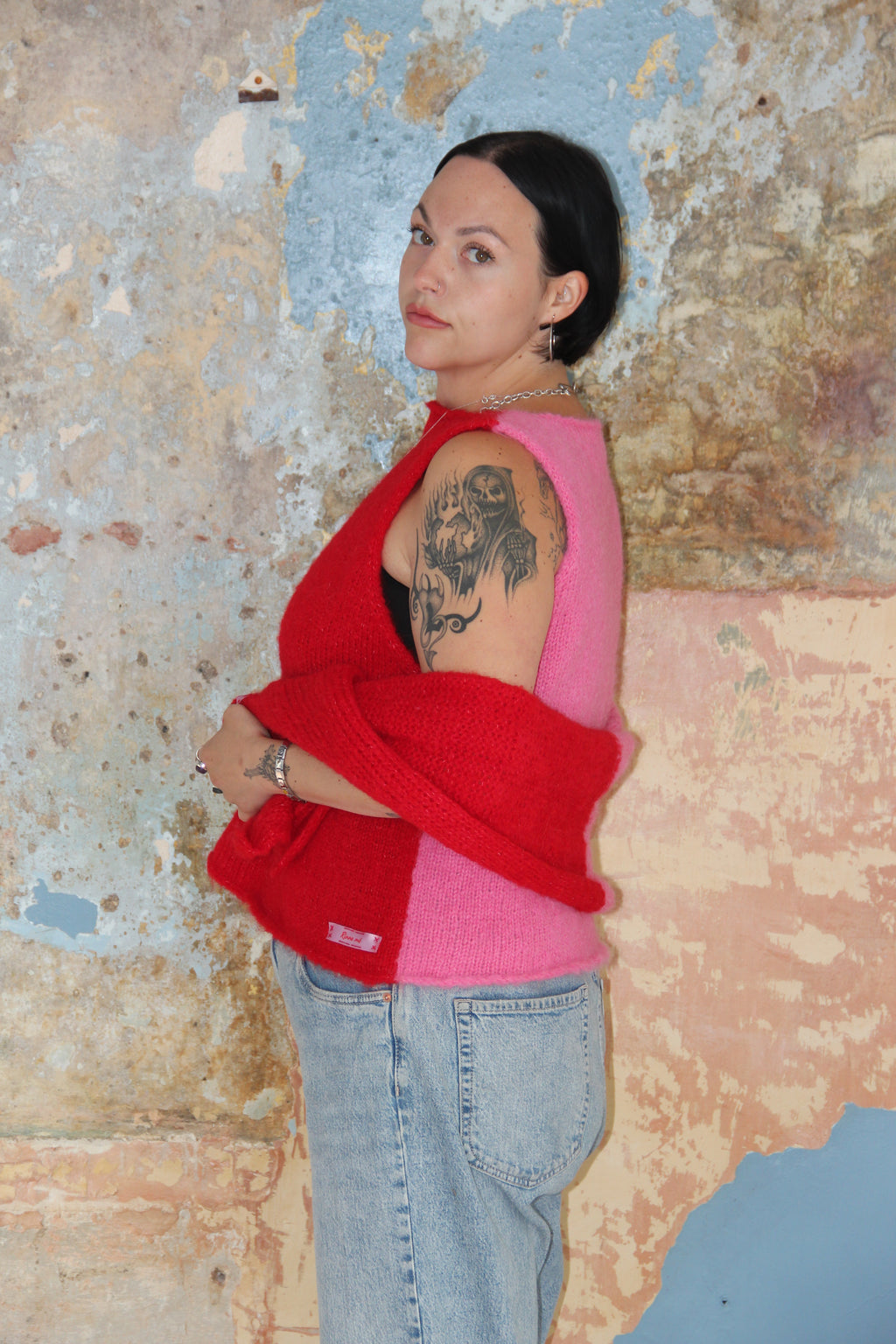 A woman with dark hair tied back stands against a textured plaster wall, wearing a red and pink colour-block knit sweater paired with light-wash blue jeans.